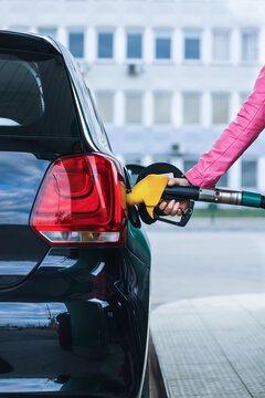 Woman holding fuel nozzle and refueling black car at gas station, closeup. Concept of fuel price, travel, car rental service, driving trip and automobile maintenance.