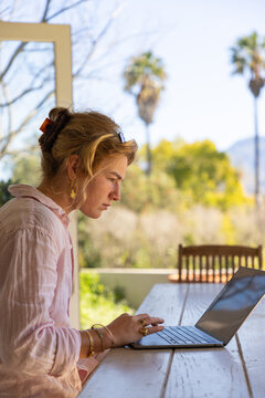 young woman typing on laptop under patio, palm trees in background