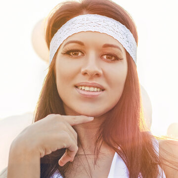 Close-up portrait of a beautiful smiling young woman with long dark hair, wearing a stylish white lace headband, touching her chin and looking at camera in warm golden sunlight outdoors.