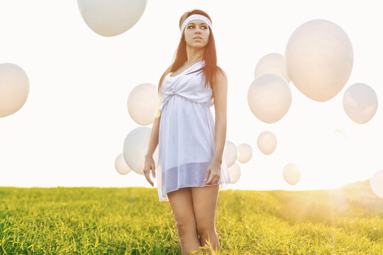 Dreamy portrait of a woman in a white dress and headband in a green grassy field with white balloons at golden hour