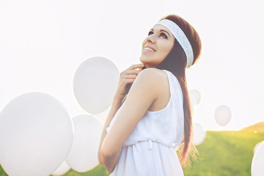 Radiant young female in a white dress and hairband looking up happily. She stands on a green meadow with floating white balloons under a warm sun. A symbol of hope, joy, and freedom.