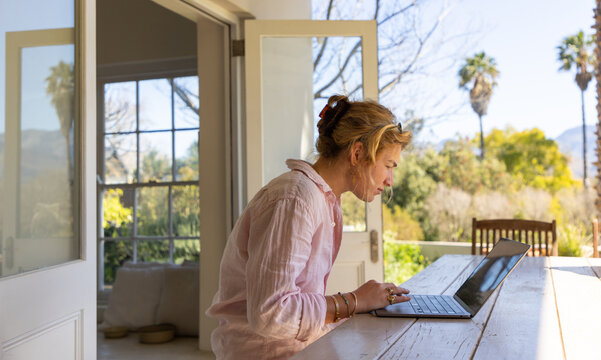 young woman typing on laptop under patio, palm trees in background