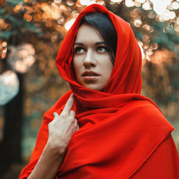 Portrait of a beautiful young woman wearing a bright red headscarf outdoors. Mysterious girl looking up against a blurred autumn forest background with golden bokeh light effects.