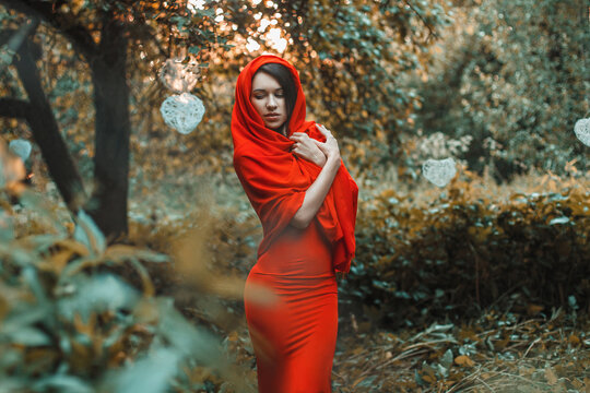 Mysterious woman in a red dress and headscarf, eyes closed, standing in a magical autumn garden with glowing white hearts. Golden hour lighting. Fine art portrait, contemplative and serene mood.