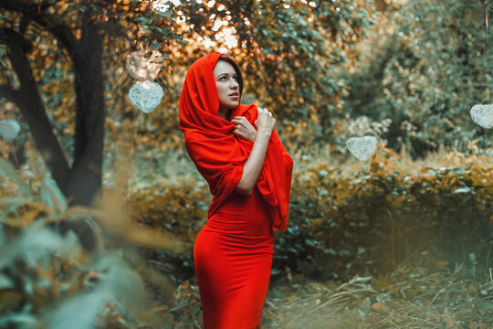 Model in tight red dress and hooded wrap standing in a natural forest clearing with hanging white wire hearts. She looks upward with a thoughtful expression. Warm sunset lighting