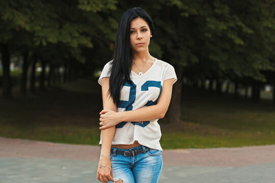 Beautiful young brunette woman with long hair wearing casual white t-shirt and jeans, standing and posing confidently in a green summer park with trees in background