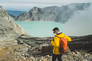 A masked Asian hiker in a yellow jacket and orange backpack stands on rocky ground overlooking a turquoise volcanic Ijen crater lake, surrounded by misty cliffs and dramatic, remote mountain scenery e © Alfian Muzaki