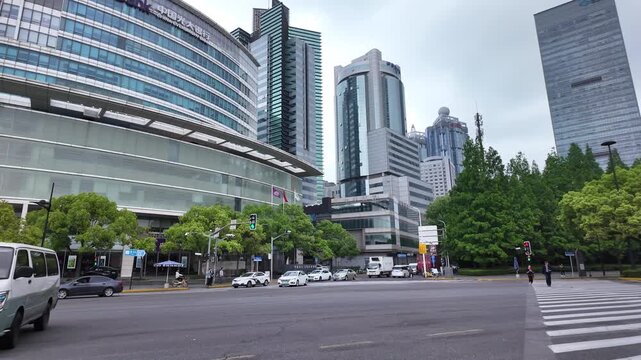 A wide urban street crossing with modern high-rise buildings and green trees under an overcast sky, with various vehicles on the road.