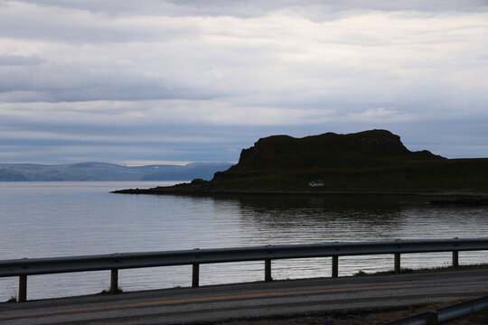 A coastal road with a guardrail overlooking a calm fjord and distant rocky hills under a cloudy sky in Norway.