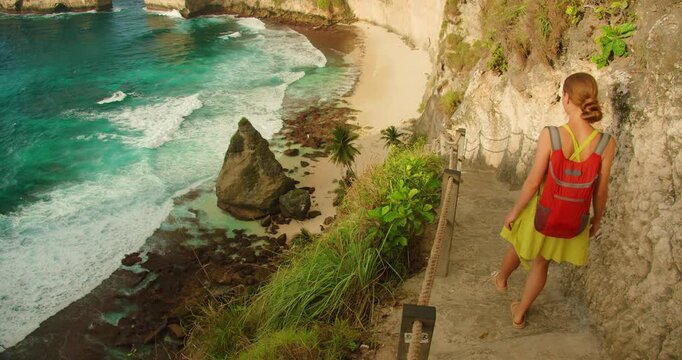 Young traveler walking along a cliffside path with a bag overlooking vast blue ocean in Nusa Penida Bali