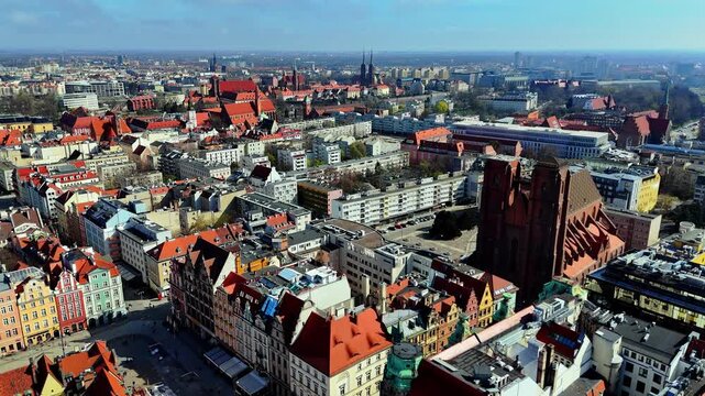 Drone footage showing an aerial panorama of Wroclaw old town, capturing historic red brick church architecture, traditional tile rooftops, Odra river, and urban cityscape under a clear blue sky in bri