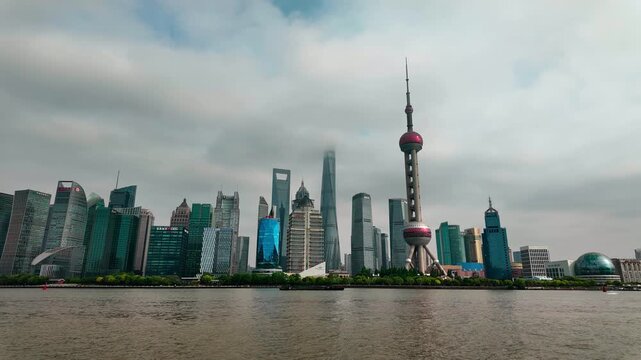 A dense modern urban skyline of tall skyscrapers lines a calm river under an overcast cloudy sky.
