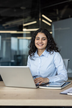 Young female professional is smiling at camera while working on her laptop at a desk in a corporate office, showing dedication and modern business environment