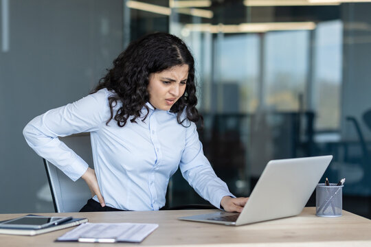 Young woman feeling severe low back pain while sitting at her office desk and working on a laptop, suffering from discomfort and health problems due to prolonged sedentary work