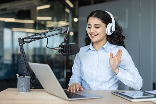Young woman creating digital audio content, speaking into a professional microphone, wearing headphones, and using a laptop during a live broadcast in a modern studio