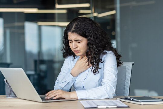 Young woman at her office desk working on a laptop, touching her neck and grimacing from a sore throat and fatigue, looking unwell and uncomfortable while trying to work