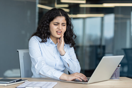 Young woman feeling sharp dental pain, holding her cheek while trying to concentrate and work on a laptop at an office desk, illustrating discomfort and health issues impacting productivity