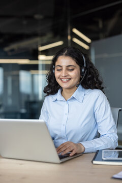 Young indian woman wearing a headset and light blue shirt, smiling and typing on a laptop while providing customer support in a modern office environment
