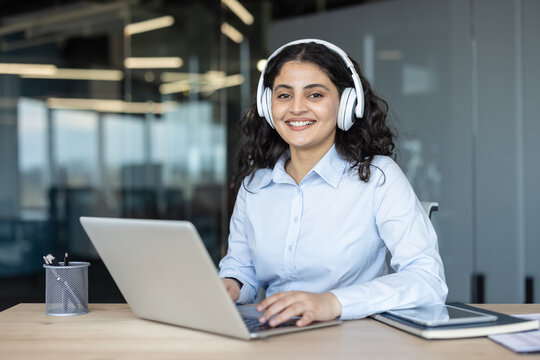 Young woman looking at camera, smiling while wearing headphones and typing on a laptop, engaged in customer support or online communication in a modern office