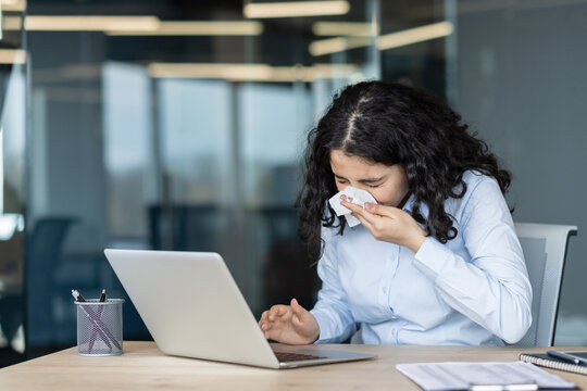 Young woman with curly hair feeling unwell, sneezing into a tissue while working on her laptop at an office desk, illustrating illness, allergies, or a common cold affecting productivity