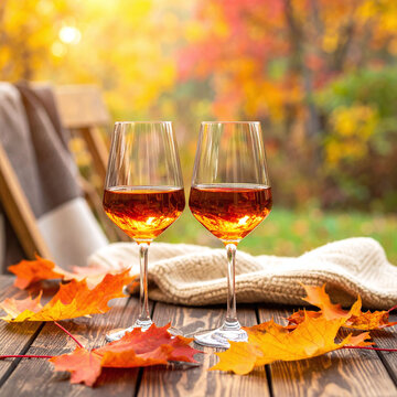 Two glasses of amber wine sit on a rustic wooden table with bright autumn maple leaves and a cozy sweater with nobody around, capturing a warm, romantic, and peaceful fall feeling.