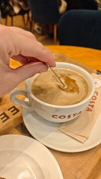 Gdansk, Poland - July 18, 2026: Hand stirring coffee in a large white cup with Costa logo on a wooden table, saucer and spoon visible in the scene