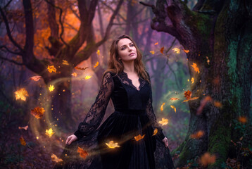 Fototapeta premium A mystical young woman in a black lace dress summoning magic leaves inside an autumn forest.
