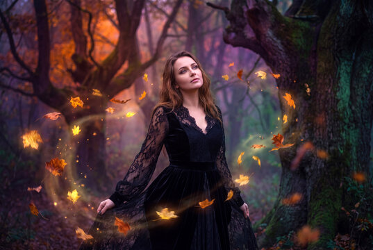 A mystical young woman in a black lace dress summoning magic leaves inside an autumn forest.