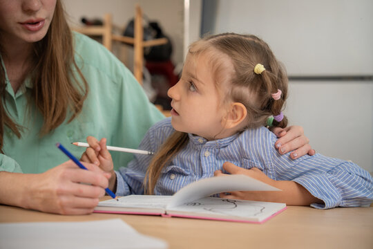 Close up of cute little girl holding pencil and talking to her mother during study session. Young girl asking her mother for help while doing primary school homework exercise at kitchen table.