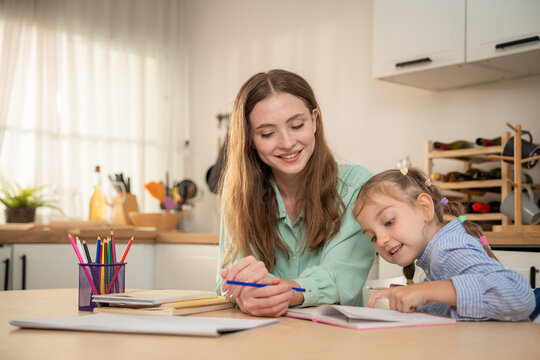 Happy mother smiling while watching her cute daughter pointing at notebook during homework. Cheerful young mother enjoying home learning time with small daughter writing and drawing at kitchen table.