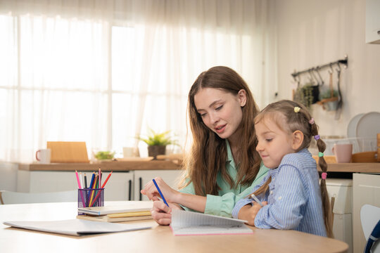 Mother helping little daughter open notebook and write homework at kitchen table. Young mother guiding her cute daughter with homeschooling study session using colored pencils in modern kitchen.