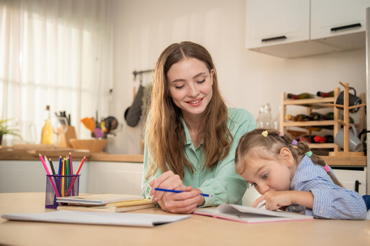 Happy mother smiling while watching her cute daughter pointing at notebook during homework. Cheerful young mother enjoying home learning time with small daughter writing and drawing at kitchen table.