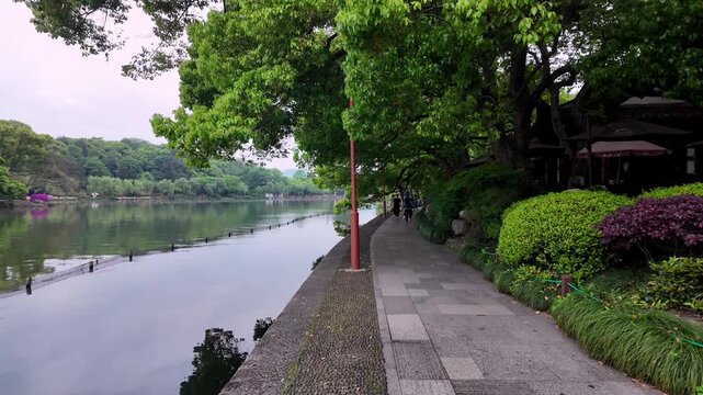 A calm lakeside paved walkway lined with lush green trees and colorful shrubs, with distant tree-covered shores under an overcast sky.