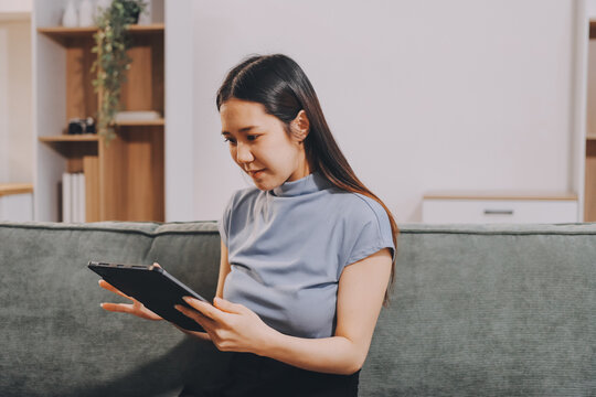 Freelance Asian woman holding mobile phone typing on laptop and working from home. Happy young girl sitting on sofa in living room.