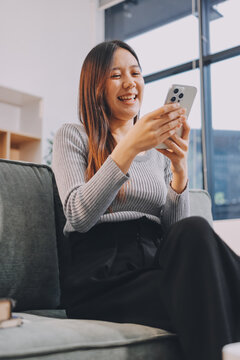 Happy relaxed young woman sitting on couch using cell phone, smiling lady laughing holding smartphone, looking at cellphone enjoying doing online ecommerce shopping in mobile apps or watching videos.