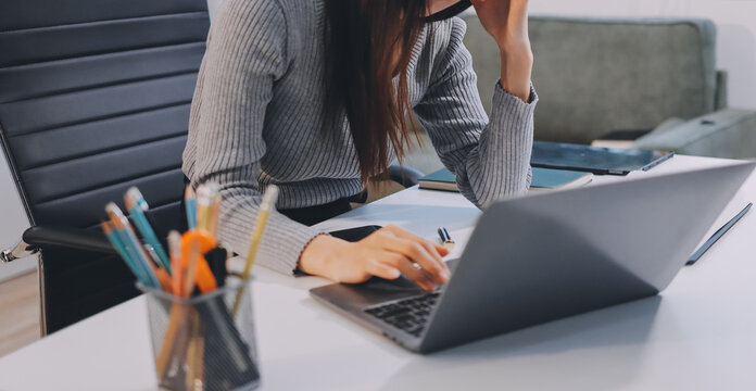 Young Asian woman sits at office desk rubbings her eyes and temples feeling pain. Stress. And exhaustion from long hours working on the computer