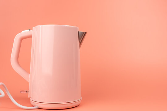 A pink electric kettle stands on a solid coral background with its spout and cord visible, leaving open space for text on the right.