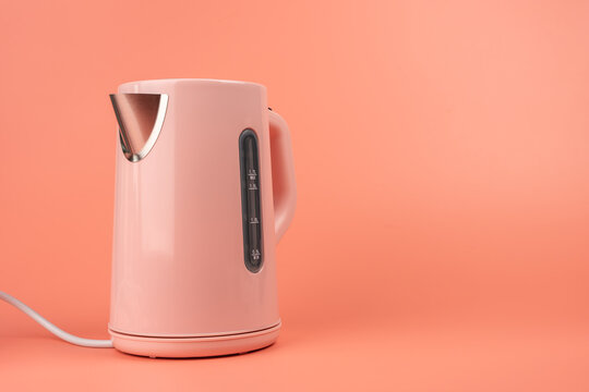 A pink electric kettle is angled to show its spout and water level window against a solid coral background with copy space.