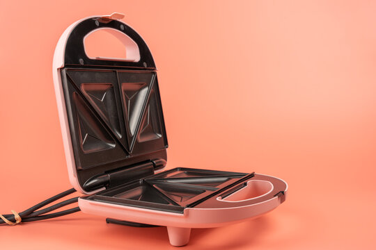 An open pink sandwich maker with black triangle plates stands alone on a solid coral background, leaving open space for text.