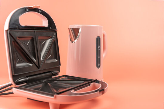 An open pink sandwich maker and a pink electric kettle are shown together at a different angle on a solid coral background.