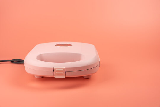 A closed pink sandwich maker is shown from a low front angle, highlighting its rounded body and rubber feet on a coral background.