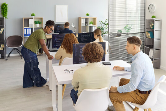 Programmers developers team coding in a busy office. Workspace shows collaboration and teamwork at computers and monitors during a mentor led meeting. Clear modern tech teamwork concept.
