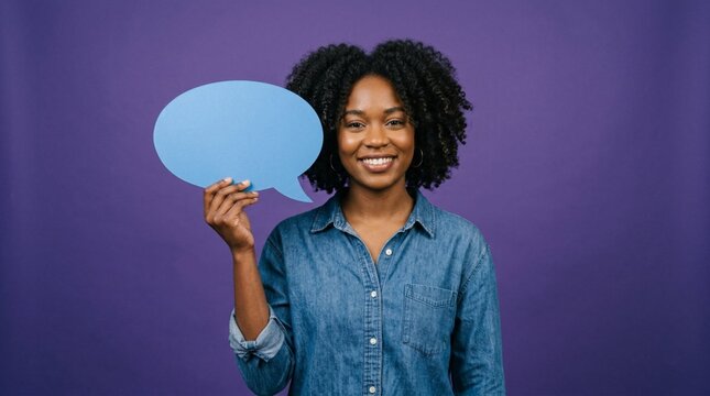Smiling young Black woman with natural curly hair holding blank blue speech bubble cutout prop against vibrant purple background, casual denim shirt portrait for communication concept