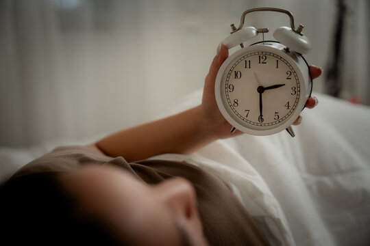 Insomnia concept of a young woman using a smartphone while lying awake in bed at night beside an alarm clock, showing sleeplessness, insomnia, smartphone, bedtime, sleep, fatigue