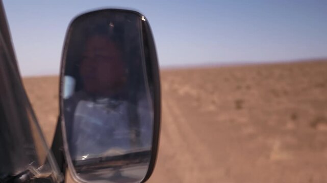 View from a car window of a side mirror reflecting a woman driver on a dusty desert road