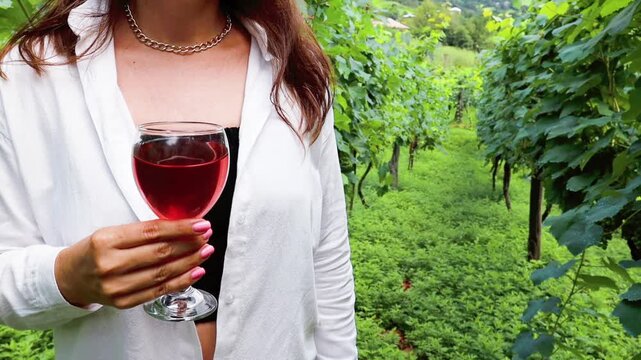 Woman unrecognized wearing the white shirt holding the glass of rose wine standing at the green vinery farn in Georgia