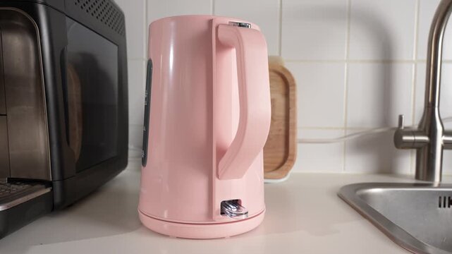 A hand sets a pink electric kettle onto its power base on a kitchen counter, then presses the power button near a sink and cutting board.