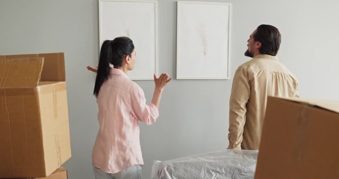 Couple moving boxes in home apartment. They hang pictures on the wall, unpacking and decorating together. Boxes and covered furniture hint at a recent move. Clear concept of move and teamwork