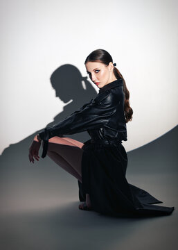 Studio portrait of gorgeous young woman sitting on a floor. Elegant cute girl dressed in black leather trench coat. Round gobo light effect. Profile view
