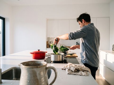 Young man cooking fresh leafy green vegetables in a pot in a modern white kitchen